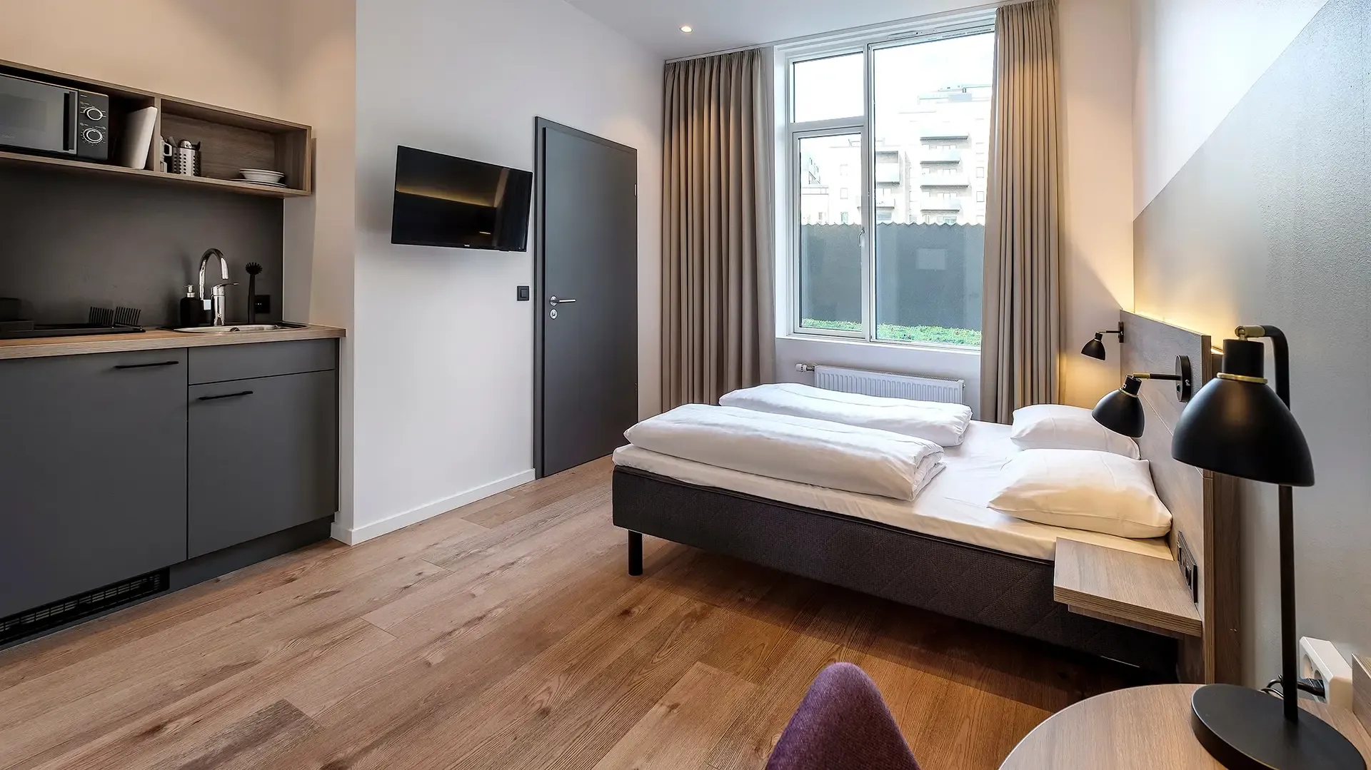Modern hotel studio room featuring kitchenette, double bed, and large window with natural light.