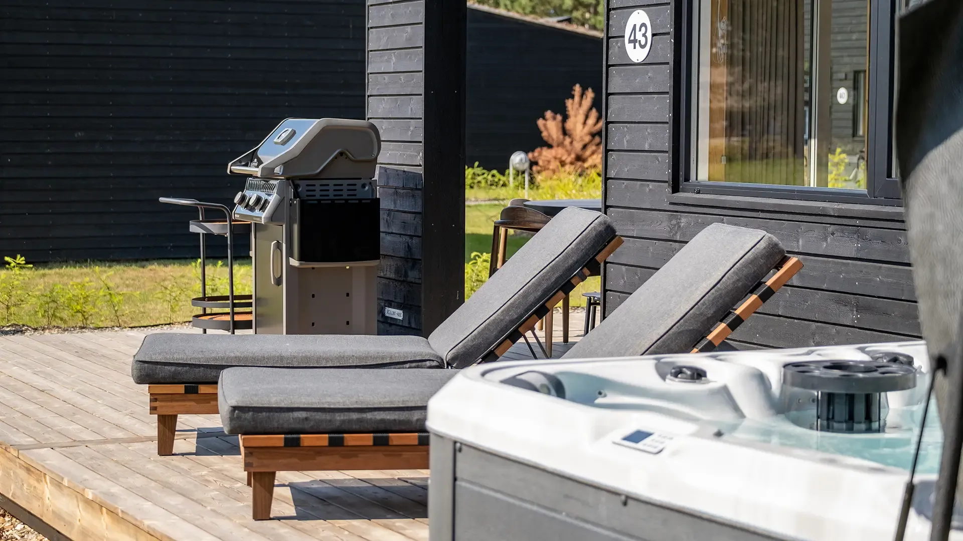 Outdoor relaxation area with sunbeds, grill and jacuzzi next to Scandinavian black wood cabin – ideal for resort guests seeking comfort and privacy.