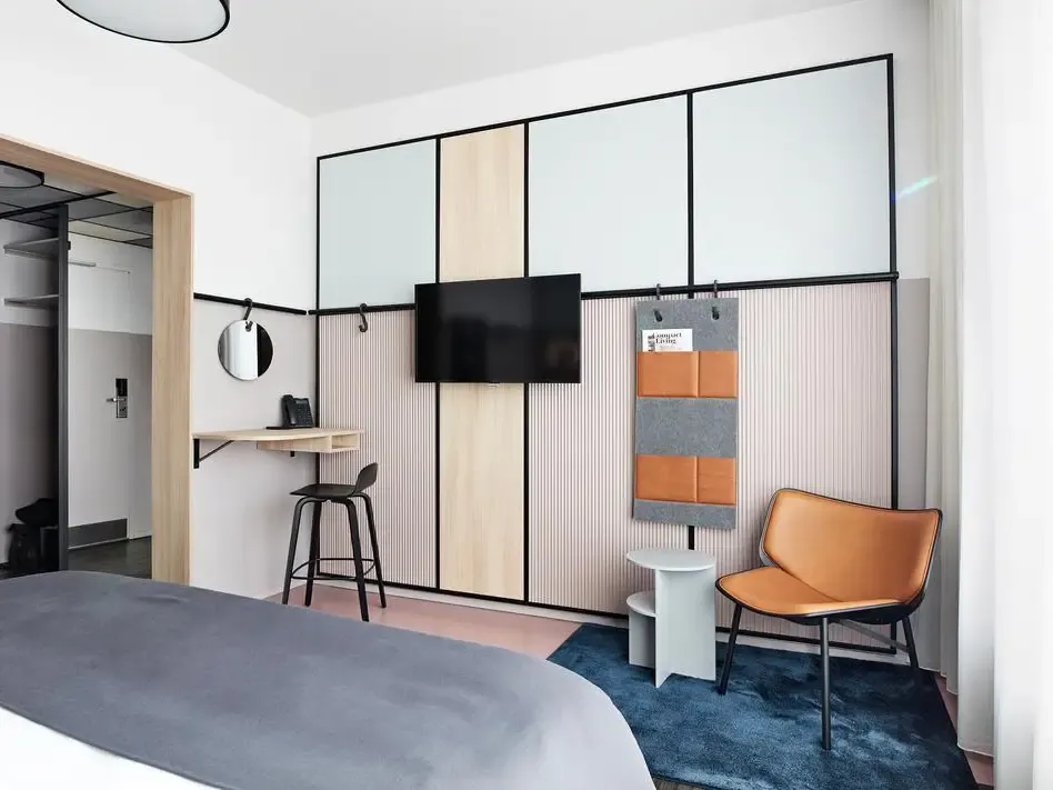 Contemporary hotel room interior featuring a cognac leather lounge chair, multifunctional wall panels, and mounted TV.