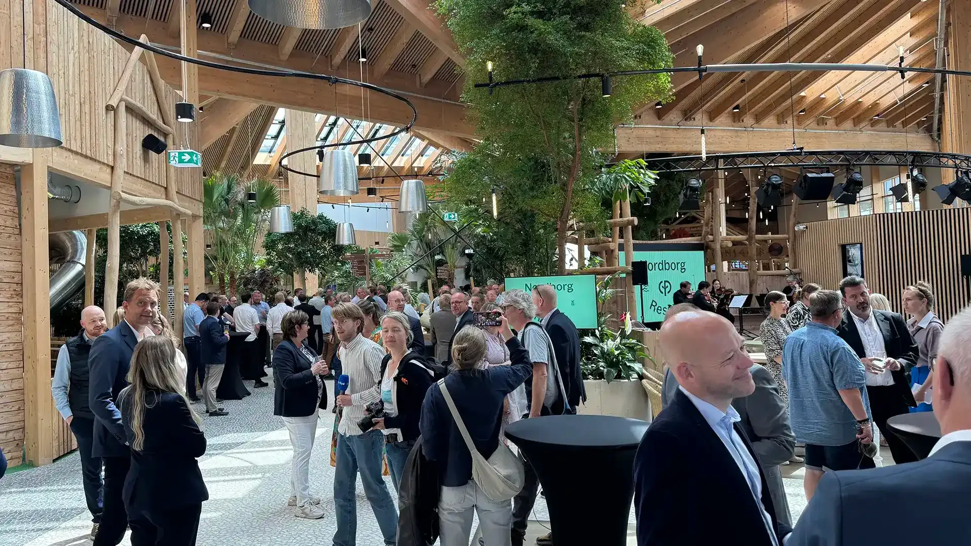 Crowds attending the Nordborg Resort opening event in the Market Dome with wooden architecture and lush greenery.