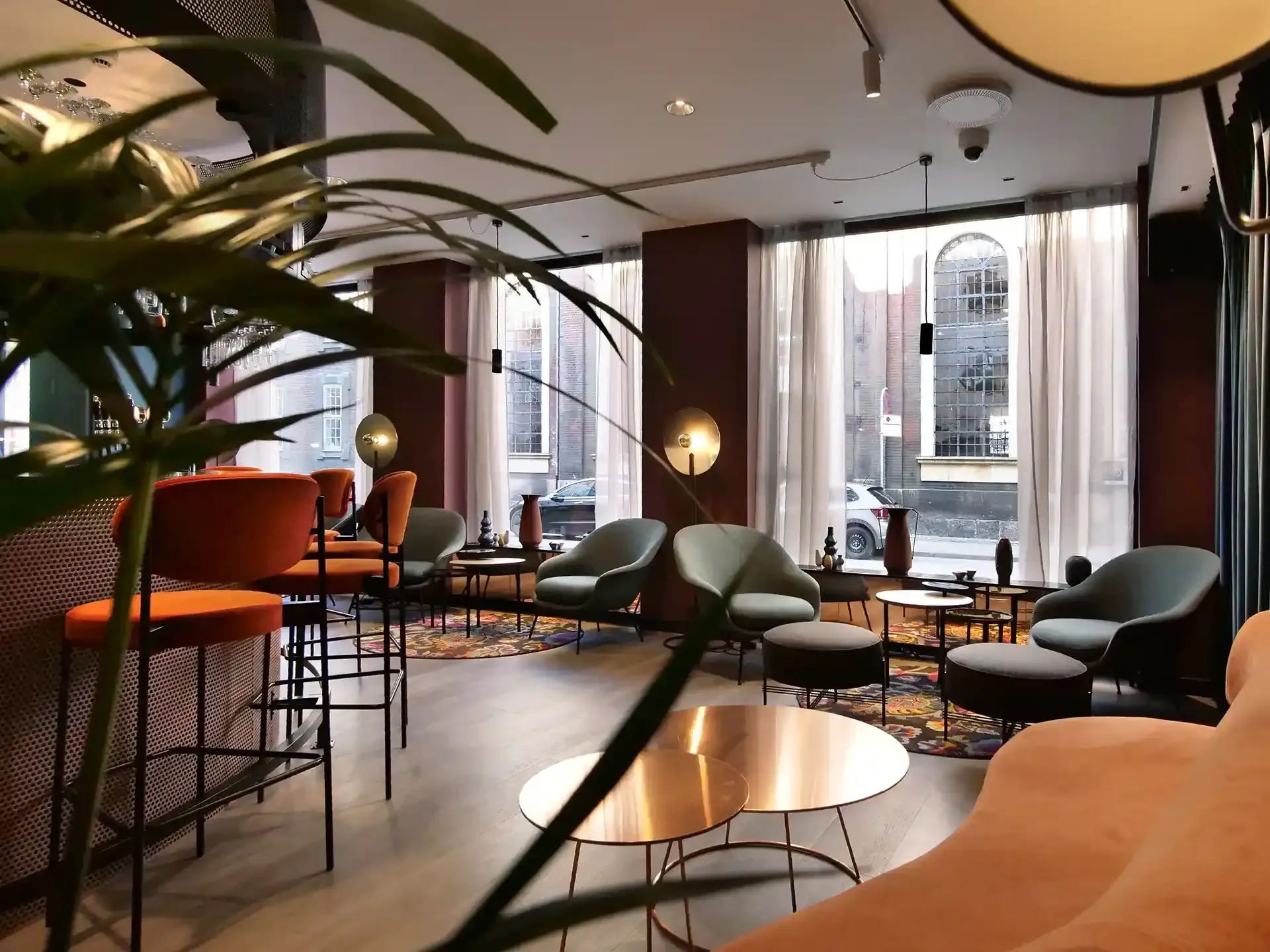 Hotel lounge seating with orange barstools, brass details and large street-facing windows – a warm and inviting space for social moments.